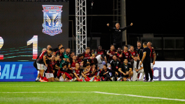 Radość piłkarzy Lincoln Red Imps FC (fot. Getty). Zdjęcie przedstawia drużynę Lincoln Red Imps FC, mistrzów Gibraltaru, świętującą historyczne zwycięstwo 2:1 nad Lechem Poznań w meczu Ligi Konferencji. Zawodnicy w czerwono-czarnych strojach są zgrupowani i uśmiechnięci na murawie boiska, a jedna osoba stoi powyżej, z rękami wzniesionymi w geście triumfu. W tle widoczny jest duży ekran z herbem Lecha Poznań (fot. Getty).