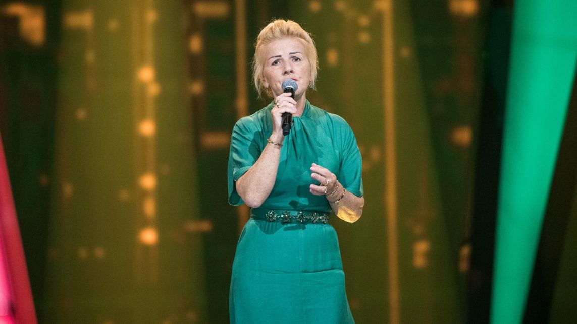 Barbara Postoł, uczestniczka programu „The Voice Senior” w butelkowozielonej kreacji, śpiewa przed trenerami piosenkę „Nie było ciebie tyle lat” Krystyny Giżowskiej.